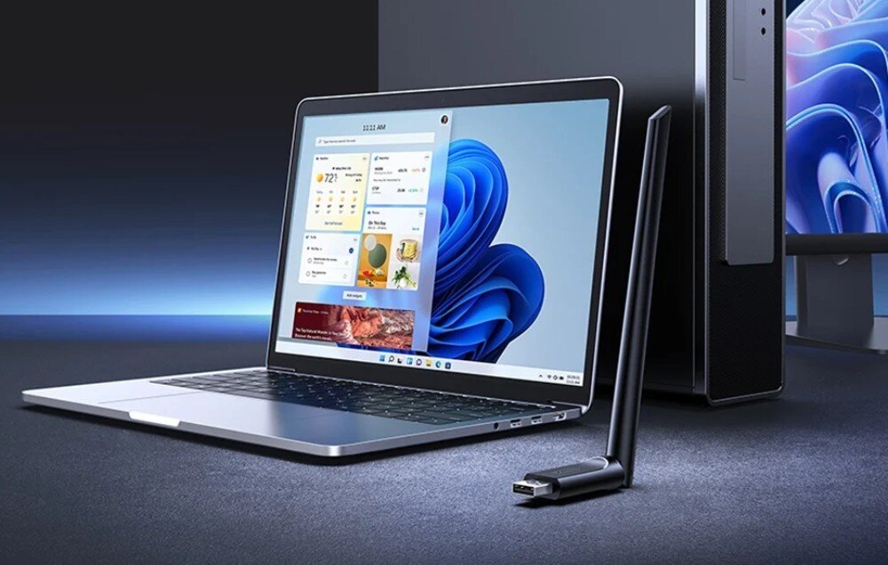 Karta sieciowa UGREEN CM763 Czarna bezprzewodowa karta sieciowa na USB z jedną długą anteną, ustawiona pionowo przed otwartym laptopem i komputerem stacjonarnym