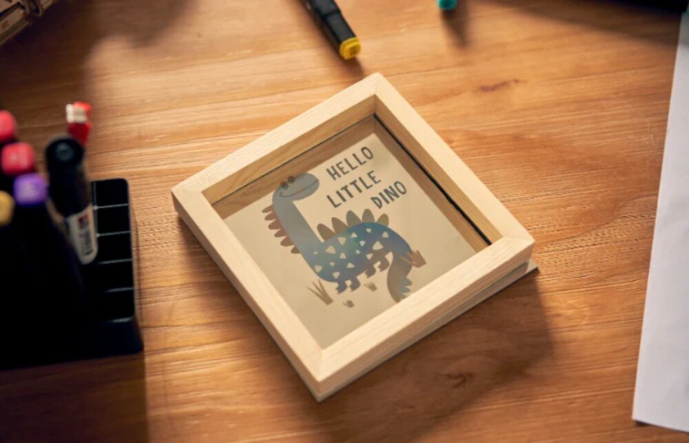 Na drewnianym stole znajduje się drewniana ramka z obrazkiem przedstawiającym niebieskiego dinozaura i napisem \'HELLO LITTLE DINO\'. Obok ramki widoczne są kolorowe markery w stojaku.