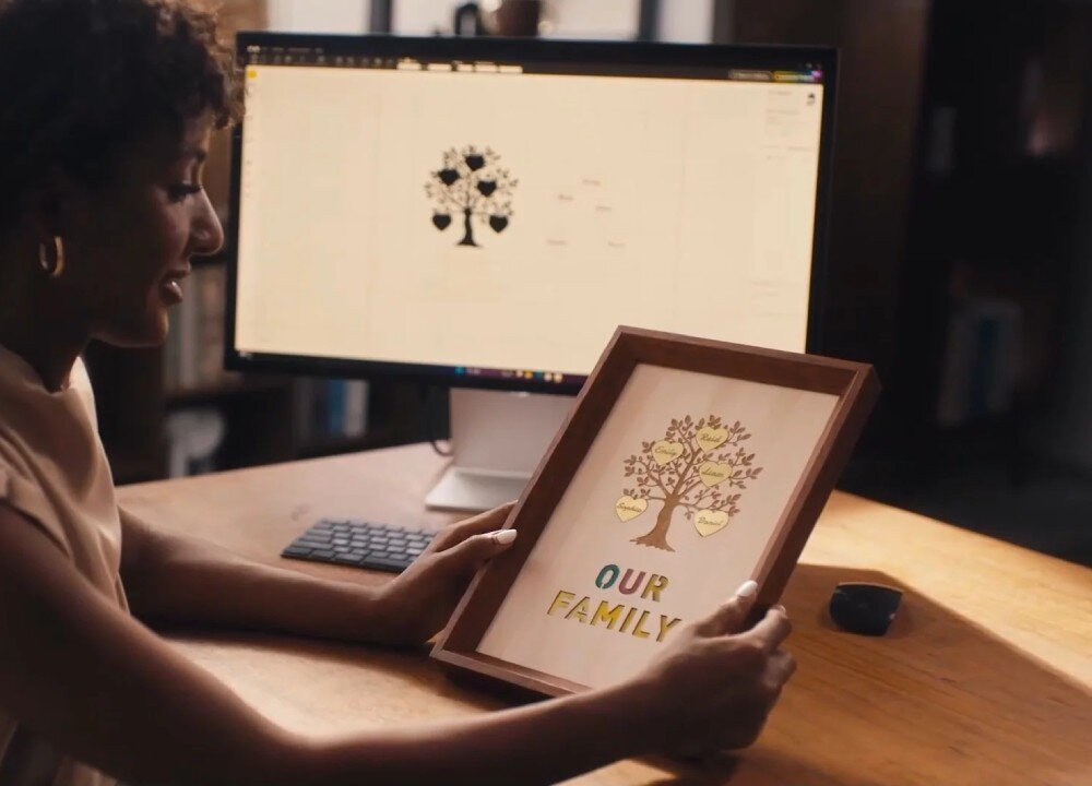 Osoba trzyma w rękach ramkę z grafiką drzewa i napisem \'OUR FAMILY\'. W tle widać monitor z podobnym projektem drzewa na ekranie.