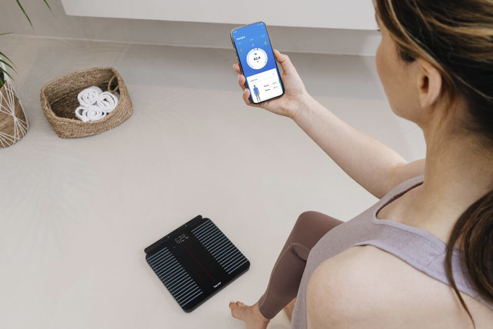 Osoba siedzi na podłodze, trzymając w ręku smartfon z aplikacją pokazującą dane dotyczące wagi. Obok na podłodze znajduje się elektroniczna waga łazienkowa z wyświetlaczem.