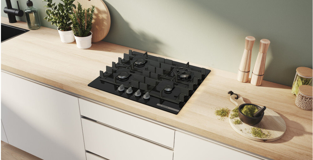 PŁYTA BOSCH PNH6B6K40 Na zdjęciu znajduje się czarna płyta gazowa umieszczona na jasnym blacie kuchennym, otoczona akcesoriami do gotowania, przyprawami oraz zielonymi ziołami w doniczkach. Całość tworzy uporządkowaną, estetyczną przestrzeń sprzyjającą przygotowywaniu posiłków