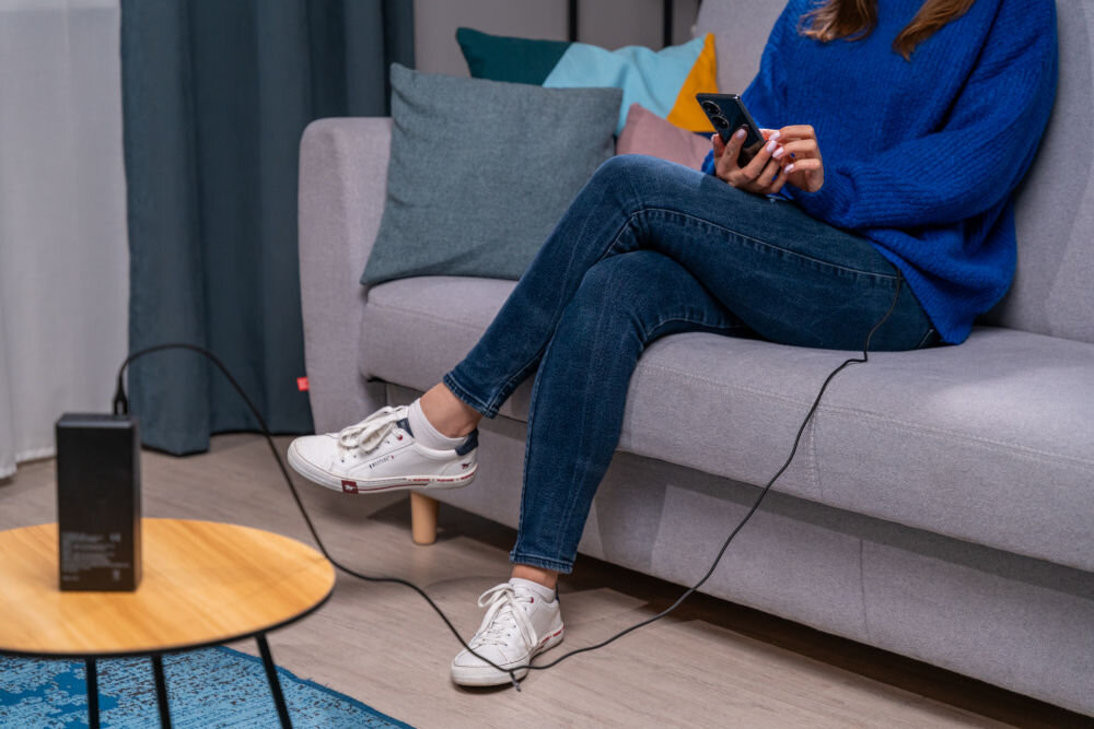 Osoba siedzi na kanapie, trzymając telefon podłączony do ładowarki za pomocą czarnego kabla USB-C. Kabel jest podłączony do urządzenia stojącego na okrągłym stoliku.