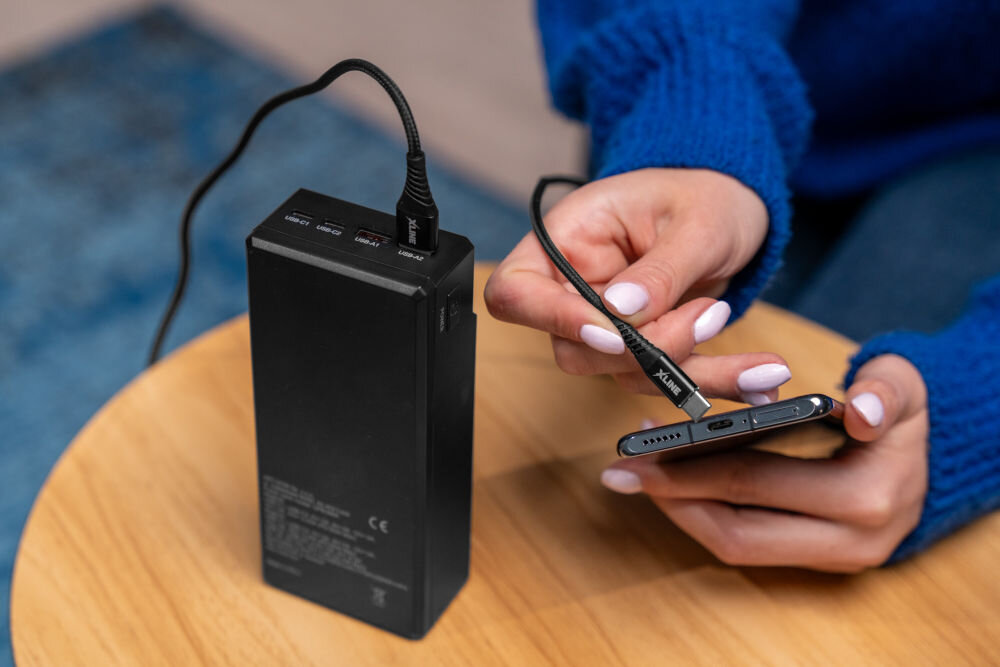 Osoba trzyma smartfon i podłącza go do czarnego powerbanku za pomocą kabla USB-C XLINE. Na kablu widoczne jest logo \'XLINE\'.
