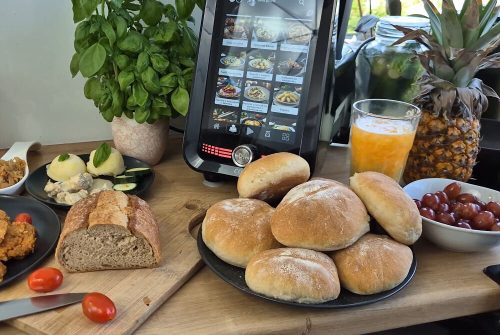 Na drewnianym blacie znajduje się robot kuchenny z ekranem dotykowym, na którym wyświetlane są przepisy kulinarne. Obok leżą bułki na talerzu, chleb, pomidorki koktajlowe, sok pomarańczowy oraz świeże zioła w doniczce.