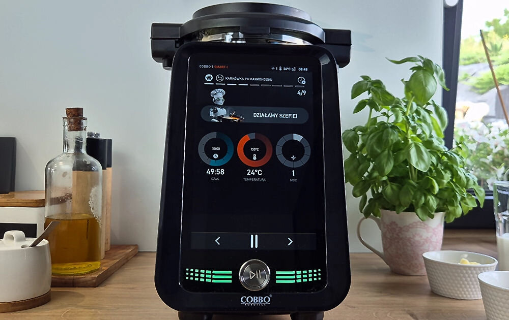 Na zdjęciu widać robot wielofunkcyjny COBBO 7 Smart-I z wyświetlaczem pokazującym czas 49:58, temperaturę 24°C i moc 1. W tle znajdują się zioła w doniczce, butelka z oliwą oraz inne kuchenne akcesoria.