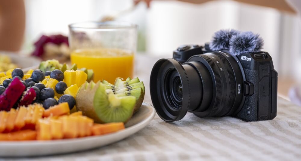 Na stole znajduje się aparat bezlusterkowy z obiektywem, obok talerza z pokrojonymi owocami i szklanki soku pomarańczowego. Na aparacie widoczne jest logo \'Nikon\'.