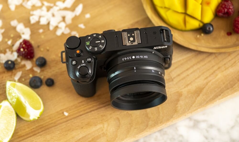 Na drewnianym stole leży czarny aparat bezlusterkowy z obiektywem, obok znajdują się kawałki limonki, jagody i plasterki mango. Na aparacie widoczne jest logo \'Nikon\'.