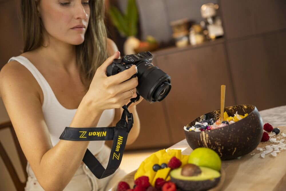 Osoba trzyma aparat fotograficzny z widocznym paskiem z napisem \'Nikon Z\'. Na stole przed nią znajduje się miska z owocami i drewnianą łyżką.
