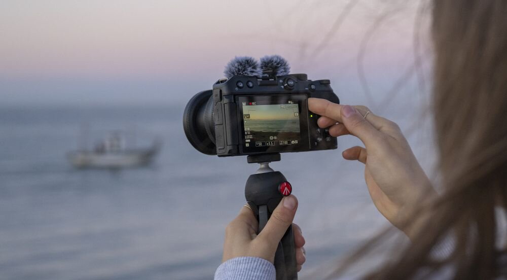Osoba trzyma aparat bezlusterkowy z mikrofonem, skierowany na morze z widoczną łodzią na ekranie. Aparat zamontowany jest na statywie z logo \'Manfrotto\'.