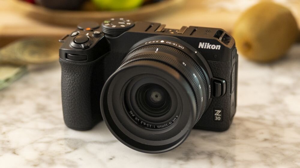 Czarny aparat bezlusterkowy z obiektywem, na którym widnieje napis \'Nikon Z 30\'. Aparat leży na marmurowym blacie, w tle widoczne są rozmyte owoce.