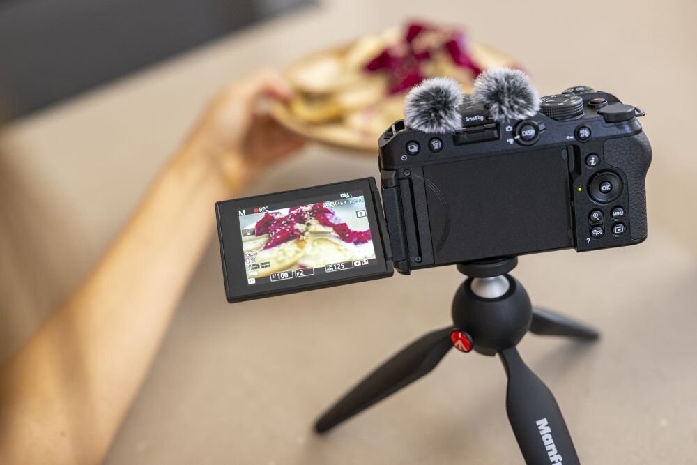 Aparat bezlusterkowy z odchylanym ekranem LCD, na którym widoczny jest obraz naleśników z owocami. Aparat zamontowany jest na statywie z logo \'Manfrotto\'.