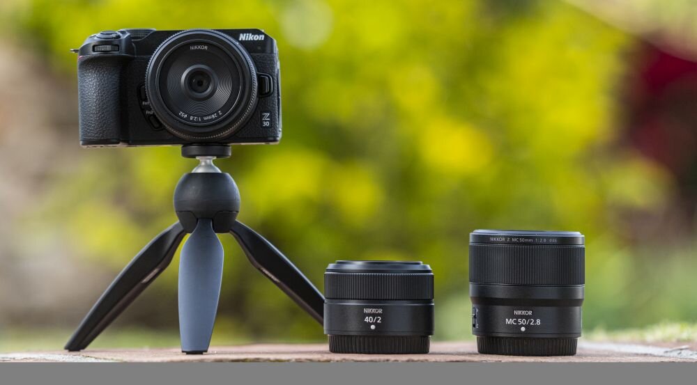 Na zdjęciu widoczny jest czarny aparat bezlusterkowy marki Nikon zamontowany na małym statywie oraz dwa obiektywy leżące obok. Na aparacie widnieje napis \'Nikon Z 30\', a na obiektywach napisy \'NIKKOR 40/2\' i \'NIKKOR MC 50/2.8\'.