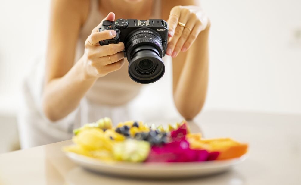Osoba trzyma aparat fotograficzny marki Nikon, robiąc zdjęcie kolorowego talerza z owocami.