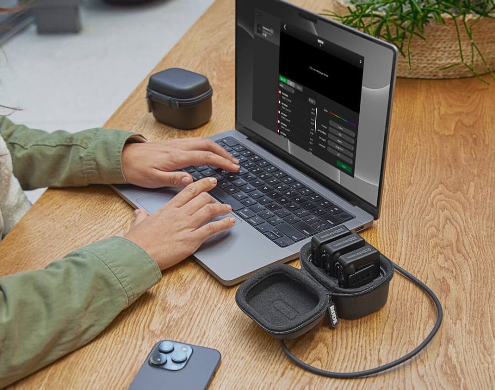 Osoba pracuje na laptopie, obok którego znajduje się system bezprzewodowy RODE Wireless Pro w etui. Na ekranie laptopa widoczny jest interfejs oprogramowania z listą ustawień.
