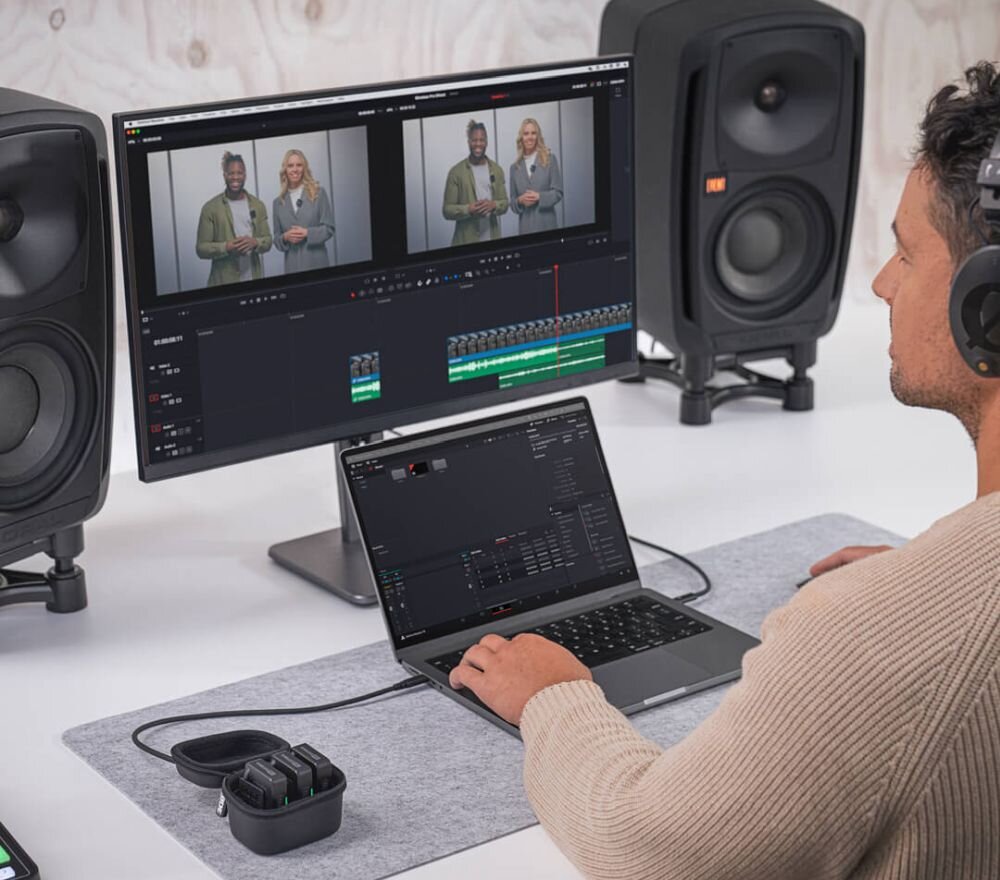 Osoba pracuje przy biurku z laptopem i dużym monitorem, na którym wyświetlane są dwa obrazy wideo. Obok laptopa znajduje się system bezprzewodowy RODE Wireless Pro.