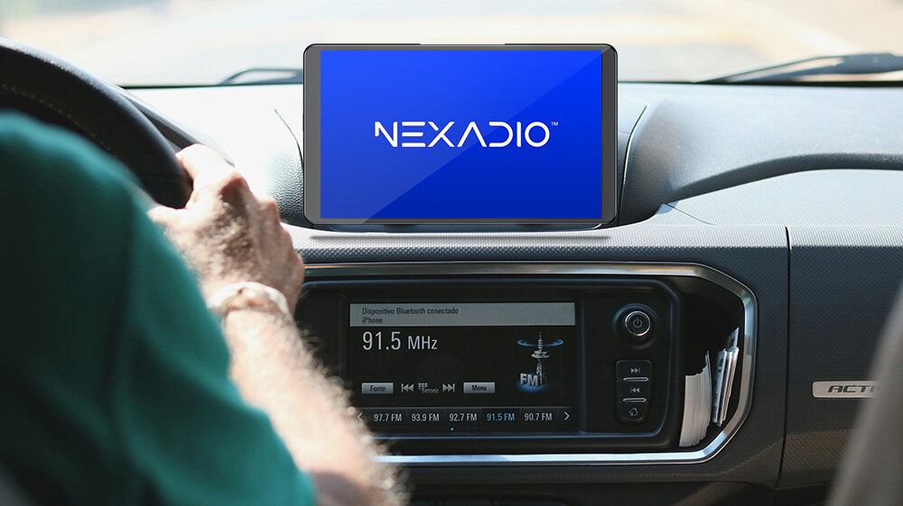 Na desce rozdzielczej samochodu znajduje się tablet z logo \'NEXADIO\' na ekranie. Poniżej widoczny jest panel radia samochodowego z wyświetlaczem pokazującym częstotliwość 91.5 MHz.