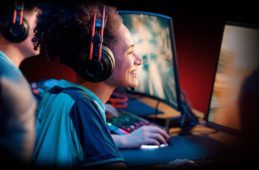 Karta graficzna XFX Swift Radeon RX 9060 XT OC Triple Fan Gaming Edition 16GB Uśmiechnięta kobieta w słuchawkach grająca na komputerze z zakrzywionym monitorem, obok druga osoba także w słuchawkach, interfejs, kompatybilność z ryzen 9000