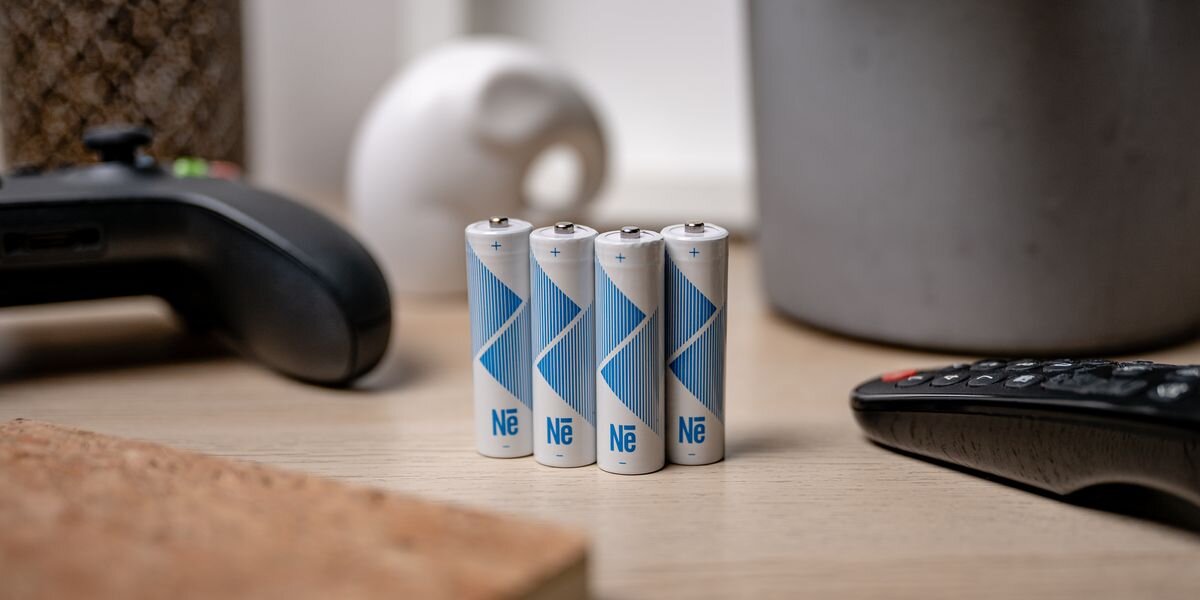 Akumulator NEWELL Li-ion AA 2200 mAh (4 szt.), Cztery białe akumulatory AA z niebieskimi detalami stoją pionowo na drewnianym stole obok kontrolera i pilota, zasilanie, dzień, pewne, moc