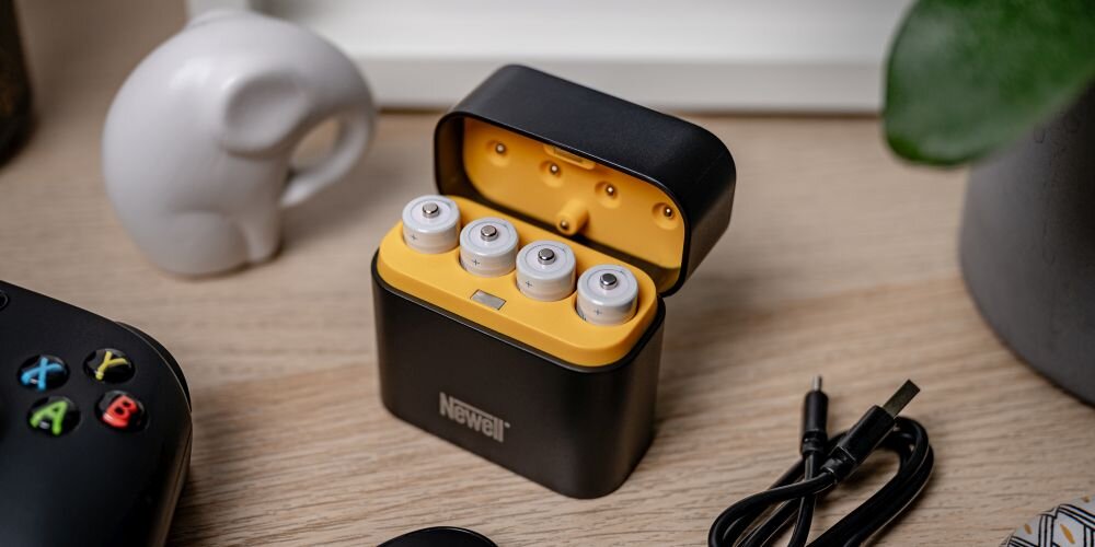 Ładowarka 4-kanałowa Newell z 4 akumulatorami Li-ion AA na drewnianym blacie. W tle porcelanowy słonik i roślina doniczkowa