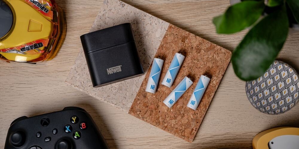 Ładowarka 4-kanałowa Newell, obok 4 akumulatory Li-ion AA na korkowej podkładce na drewnianym blacie. Wokół różne przedmioty