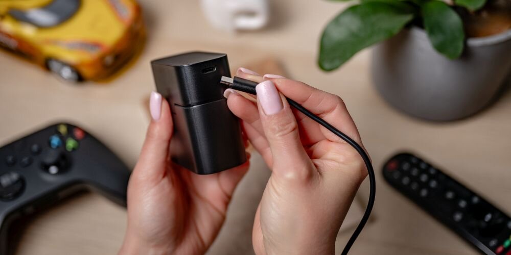 Kobiece dłonie podłączają kabel USB-C do 4-kanałowej ładowarki Newell nad drewnianym stołem, z różnymi przedmiotami w tle