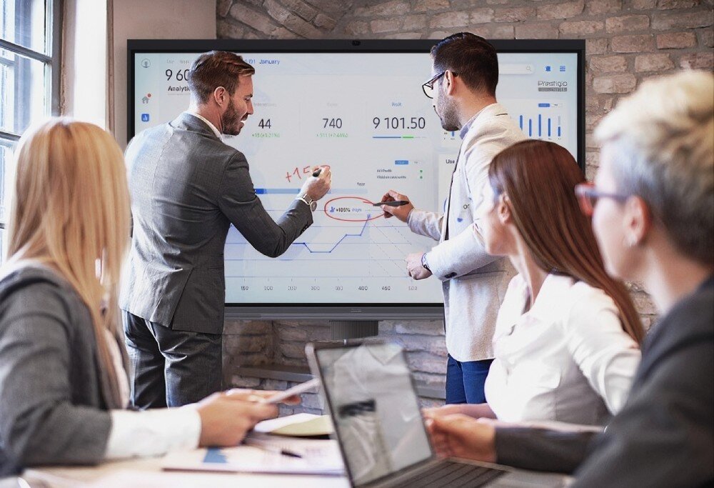 Monitor PRESTIGIO SOLUTIONS PSMB068P860 grupa osób w strojach biznesowych uczestniczy w spotkaniu, podczas którego dwóch mężczyzn nanosi notatki i zaznaczenia na wykresy prezentowane na dużym monitorze interaktywnym, ekran, przekątna, rozdzielczość