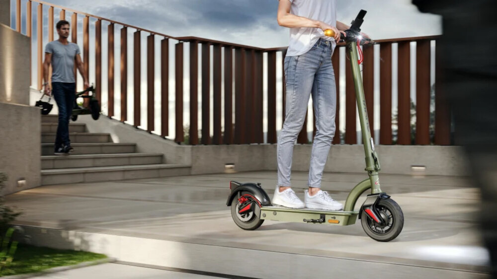 Hulajnoga elektryczna SHARP EM-KT4CEU-G 35km 350W Amortyzacja 10 cali Oliwkowo-szary miasto chodnik most jedna osoba jedzie na hulajnodze druga schodzi ze schodów z hulajnogą i kaskiem w dłoni w tle niebo barierka środek transportu styl stabilność wygodna bezpieczeństwo