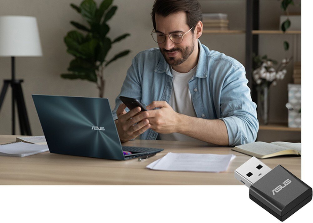 Karta sieciowa ASUS USB-BE92 Mężczyzna w okularach i niebieskiej koszuli siedzący przy biurku z laptopem oraz telefonem, obok którego widoczna jest grafika małego czarnego adaptera USB