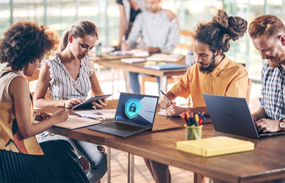 Karta sieciowa ASUS USB-BE92 Grupa osób siedząca przy drewnianym stole z laptopami, gdzie na jednym z ekranów widnieje grafika tarczy z zamkniętą kłódką