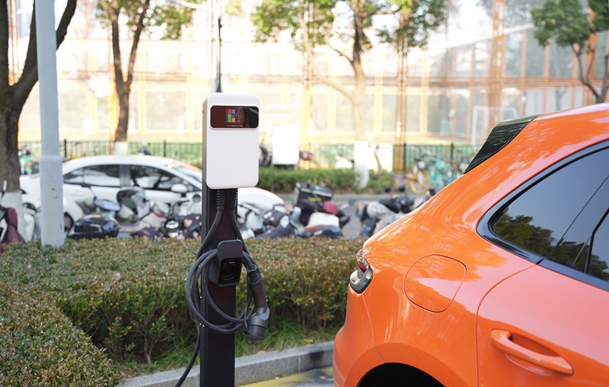 Ładowarka do samochodu elektrycznego BESEN SQ20 App 22 kW Stacja ładowania samochodów elektrycznych na słupku z zawieszonym kablem stojąca na parkingu obok pomarańczowego auta.