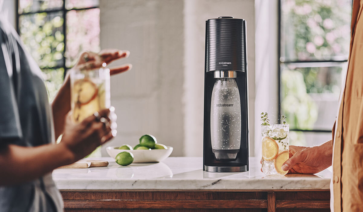 Na blacie kuchennym stoi czarny saturator z logo \'sodastream\', obok którego znajdują się szklanki z napojem i plasterkami cytryny oraz miska z limonkami.