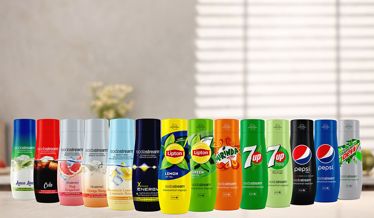 Na blacie ustawiono różnokolorowe butelki z napojami marki sodastream, w tym smaki takie jak Cola, Lipton, 7up, Pepsi i Mountain Dew. W tle widoczne są rozmyte elementy wnętrza, takie jak rośliny i okno z żaluzjami.
