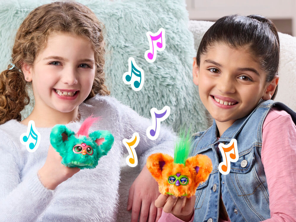 Maskotka HASBRO Furby Furbisie Boo-Kin G14355D0 Dwie uśmiechnięte dziewczynki prezentują interaktywne maskotki Furby Furblets – turkusową i pomarańczową. Wokół nich unoszą się kolorowe grafiki nutek, sugerujące funkcje muzyczne zabawek.