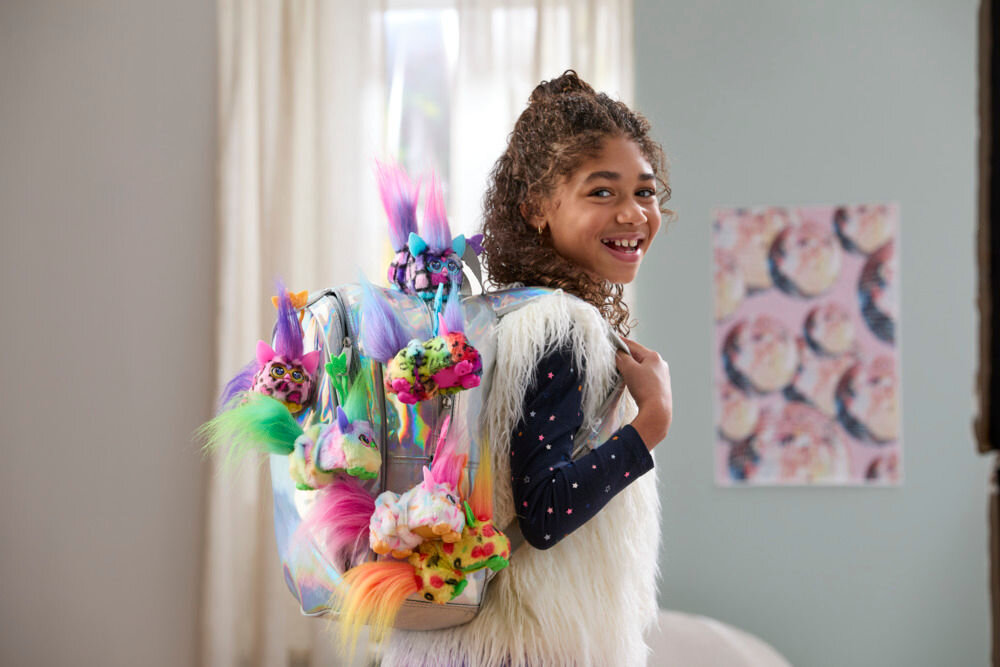 Maskotka HASBRO Furby DJ Furblets Dis-Co-Dazz G17805X0 Uśmiechnięta dziewczynka prezentuje srebrny, błyszczący plecak z przypiętą kolekcją różnokolorowych breloków Furby Furblets.