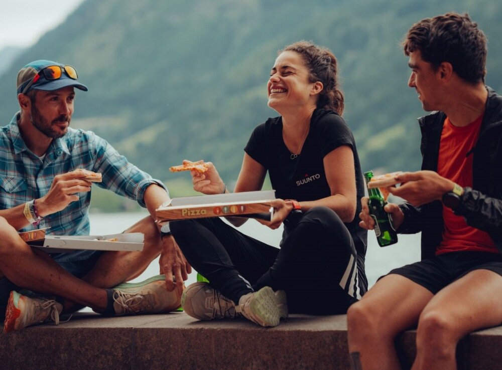 Trzy osoby siedzą na zewnątrz, jedząc pizzę z pudełek oznaczonych napisem \'Pizza\'. Jedna z osób ma na sobie koszulkę z napisem \'SUUNTO\'.