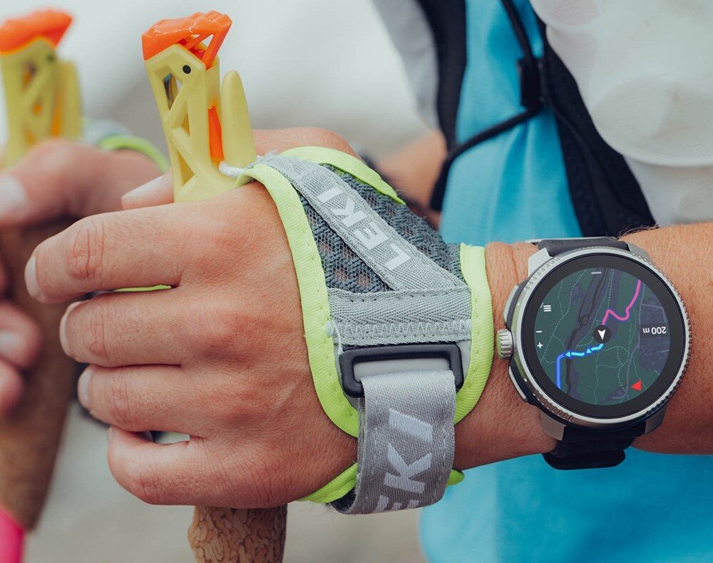 Osoba trzyma kijki trekkingowe z paskami z napisem \'LEKI\', na nadgarstku ma smartwatch z wyświetloną mapą i trasą. Na ekranie zegarka widoczna jest skala \'200 m\'.