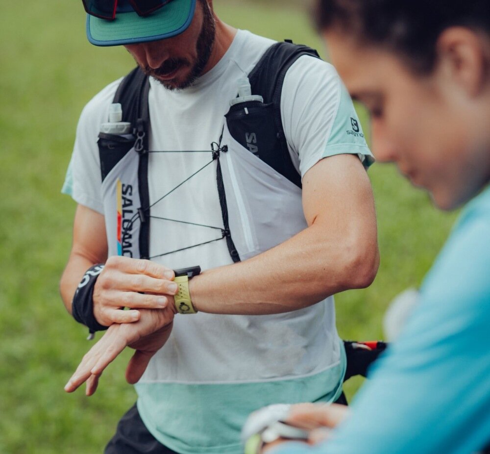 Osoba w sportowym stroju patrzy na smartwatch na nadgarstku. W tle widoczna jest inna osoba, a całość odbywa się na zewnątrz, na trawie.