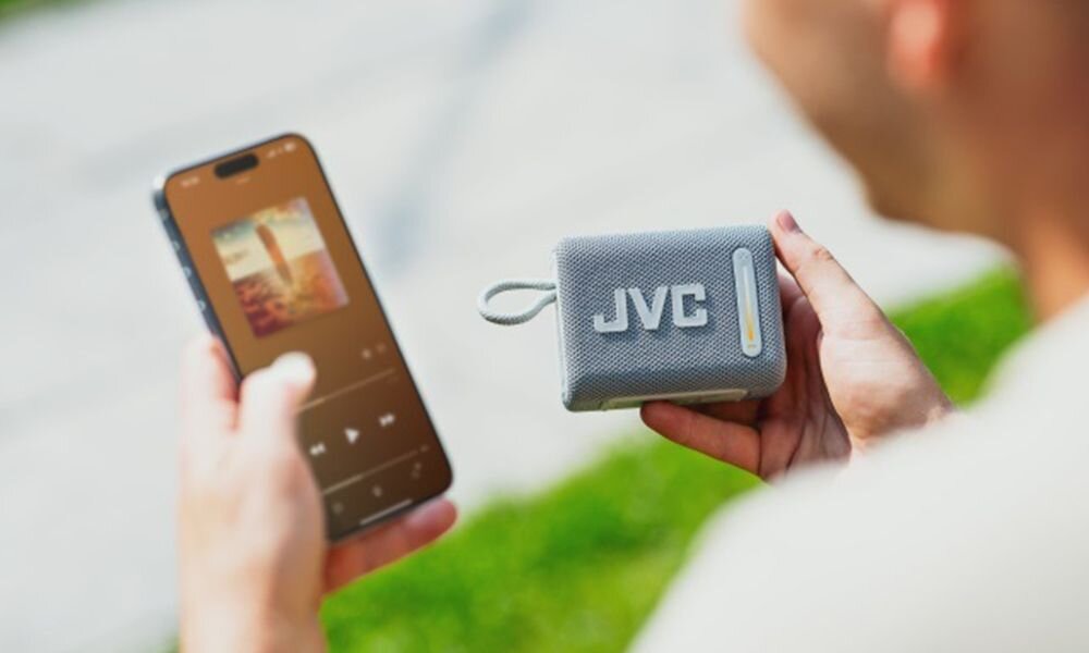 Głośnik mobilny JVC XS-E114G 5 W RMS radio FM USB MP3 pasmo 140 Hz 18 kHz, osoba trzymająca głośnik i smartfon w ręku
