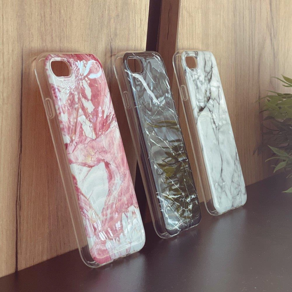 Trzy żelowe etui na telefon z marmurowym wzorem w kolorach różowym, czarnym i białym, ustawione pionowo na drewnianej powierzchni. W tle widoczna jest roślinność.