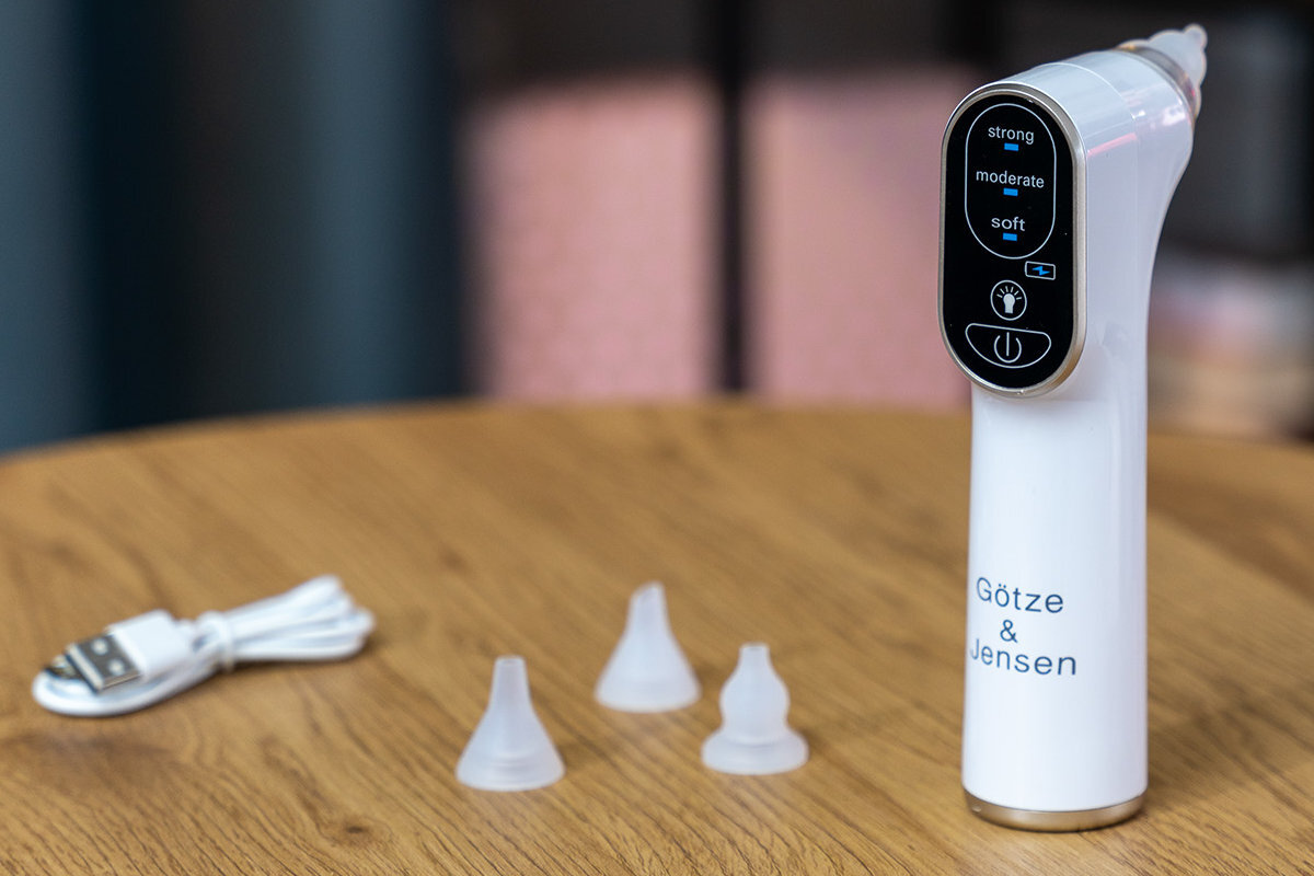 Na drewnianym stole znajduje się biały aspirator do nosa z wyświetlaczem pokazującym tryby: strong, moderate, soft. Obok leżą trzy silikonowe końcówki i biały kabel USB, na urządzeniu widnieje napis \'Götze & Jensen\'.