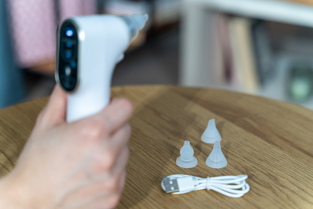 Na drewnianym stole leży biały aspirator do nosa z trzema silikonowymi końcówkami i kablem USB. W tle widoczna jest rozmyta przestrzeń domowa.