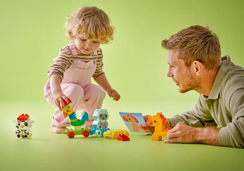 Dziecko i dorosły bawią się zestawem klocków LEGO DUPLO, przedstawiającym zwierzątka na zielonym tle. Widoczne są kolorowe klocki w kształcie zwierząt, w tym lew i papuga.