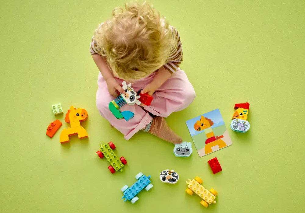 Dziecko siedzi na zielonej podłodze, bawiąc się kolorowymi klockami LEGO DUPLO w kształcie zwierząt. Obok znajdują się różne elementy konstrukcyjne i obrazek z rysunkiem zwierzątka.