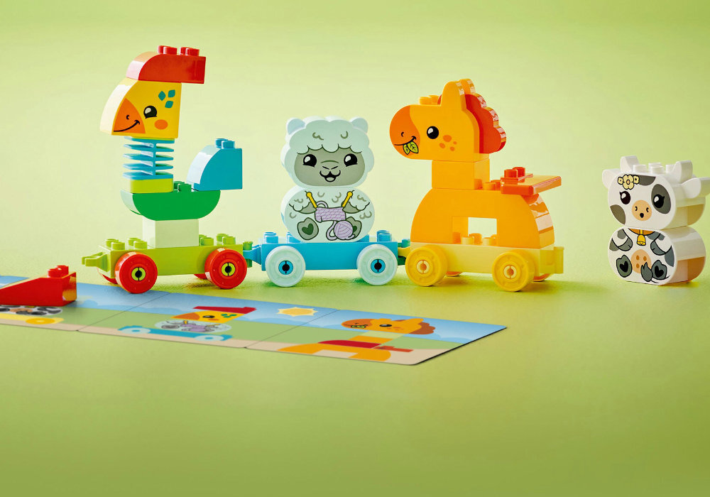 Na zielonym tle znajduje się zestaw klocków LEGO przedstawiający kolorowy pociąg z wagonikami w kształcie zwierząt. Obok pociągu leży kartonowa karta z ilustracjami zwierząt.