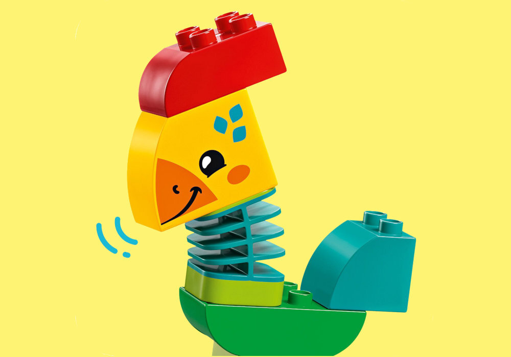 Kolorowa zabawka z klocków LEGO przedstawiająca ptaka z czerwonym grzebieniem, żółtą głową z uśmiechniętym dziobem i niebieskimi ozdobami na tle żółtego. Ptak ma zielono-niebieskie ciało z elementami przypominającymi sprężyny.