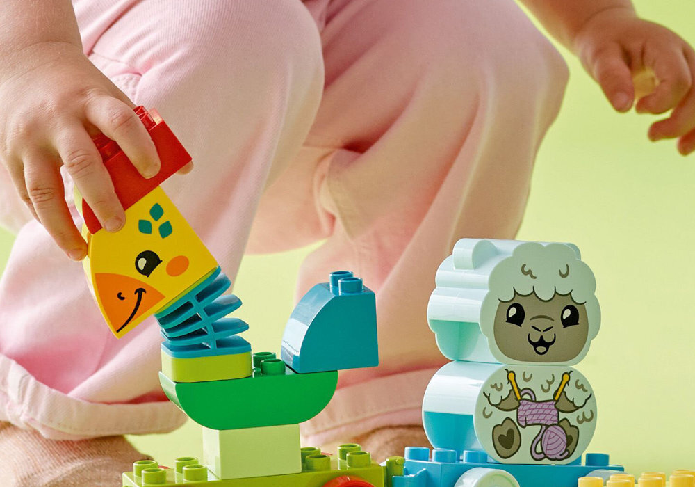 Na obrazie widać kolorowe klocki LEGO DUPLO przedstawiające zwierzątka, w tym żółtą żyrafę i białą owcę. Dziecko w różowych spodniach bawi się klockami, trzymając jeden z nich w ręce.