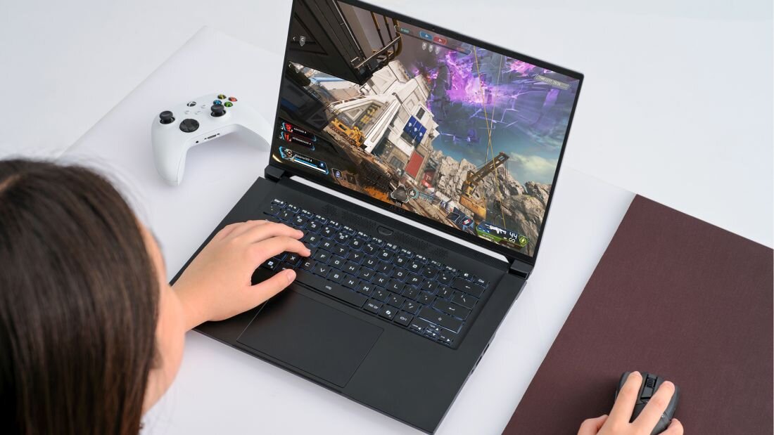 Osoba używa laptopa z wyświetlonym na ekranie dynamicznym obrazem z gry. Obok laptopa znajduje się biały kontroler do gier i myszka na podkładce.