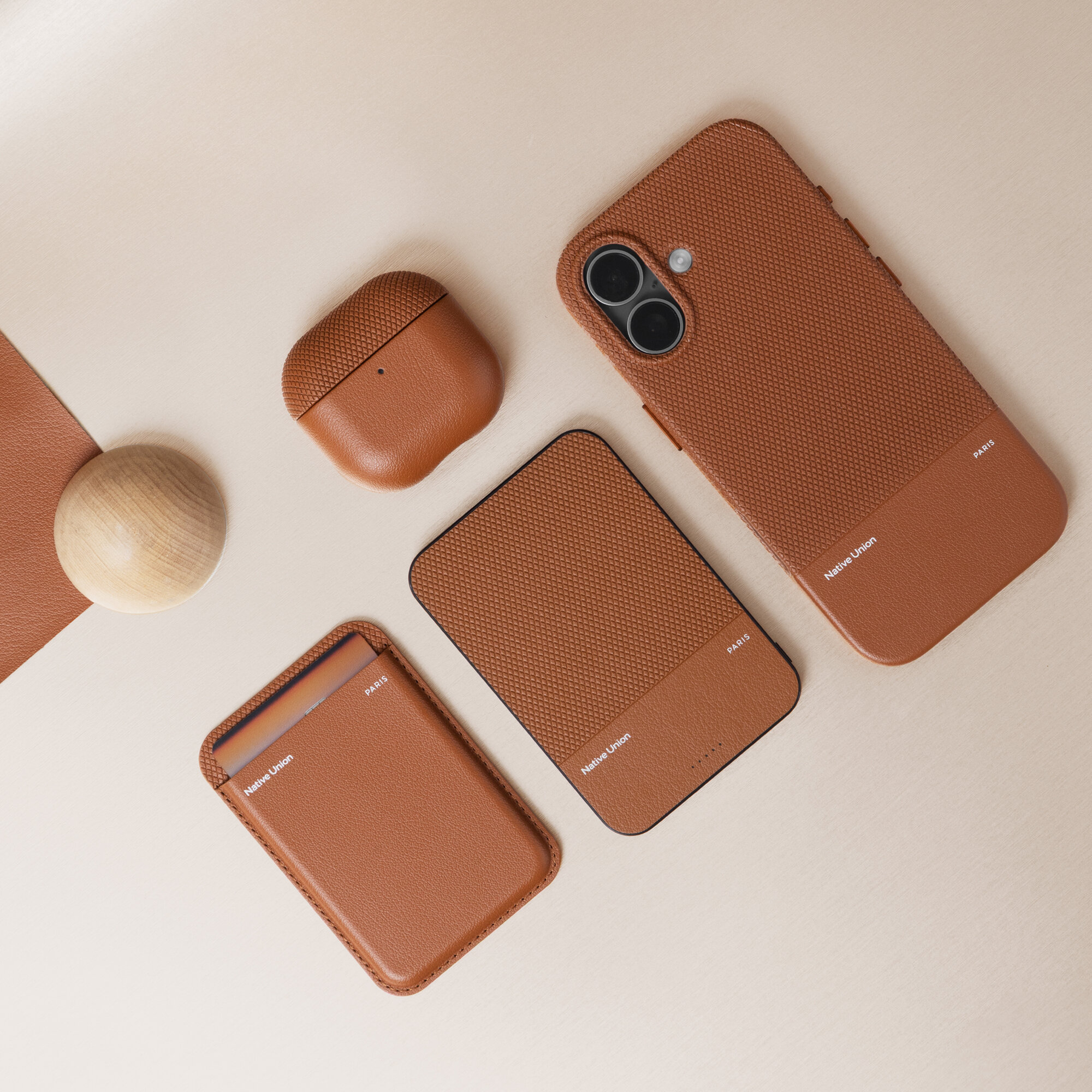 Na beżowym tle znajdują się cztery produkty w kolorze brązowym, w tym etui na telefon, etui na słuchawki, power bank oraz portfel. Na produktach widoczny jest napis \'NATIVE UNION\'.