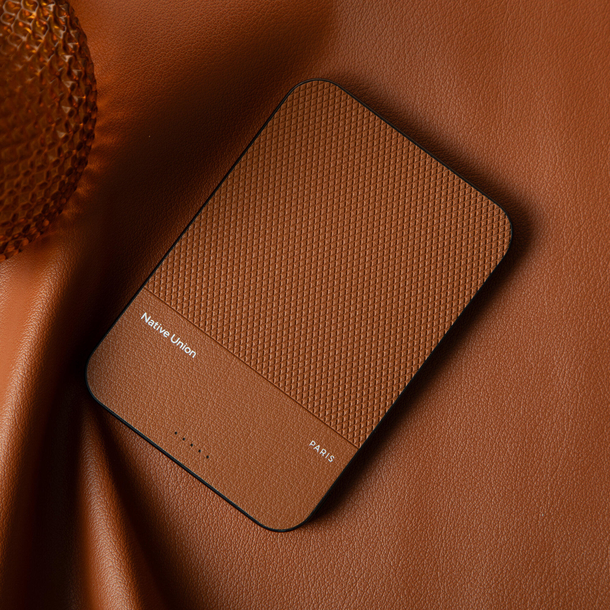 Na brązowym tle leży prostokątny, brązowy power bank z teksturą. Widoczny jest napis \'Native Union\' oraz \'PARIS\'.