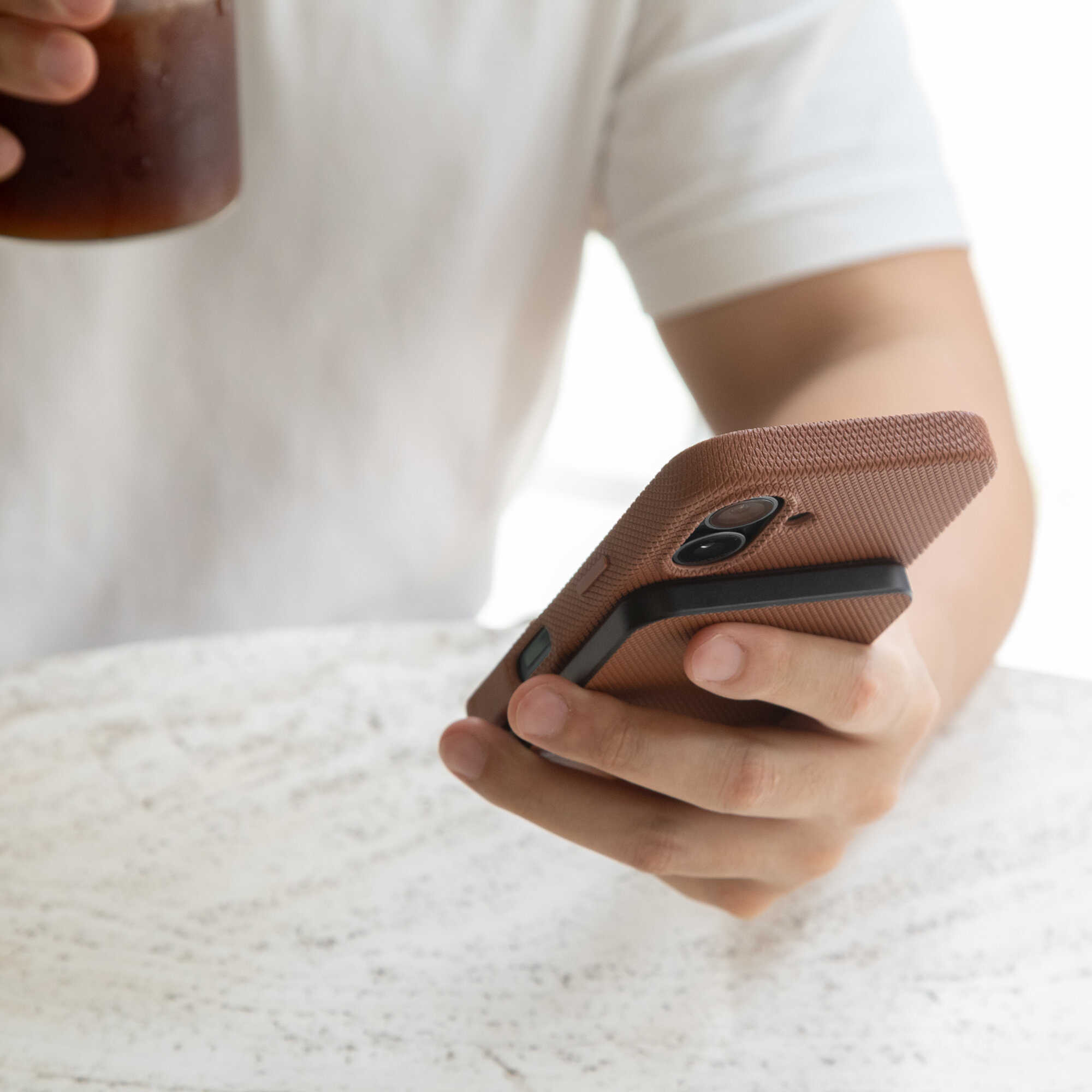 Osoba trzyma telefon z brązowym etui i przymocowanym power bankiem, siedząc przy marmurowym stole i pijąc napój z kubka.